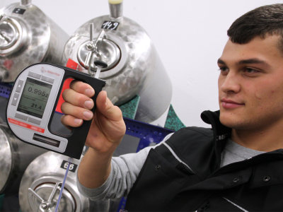 Ein Studierender mit einem Messinstrument in der Hand mit Edelstahltanks im Hintergrund